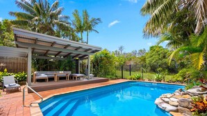 Uma piscina com deck de madeira, cadeiras de espreguiçadeira e área de estar coberta.