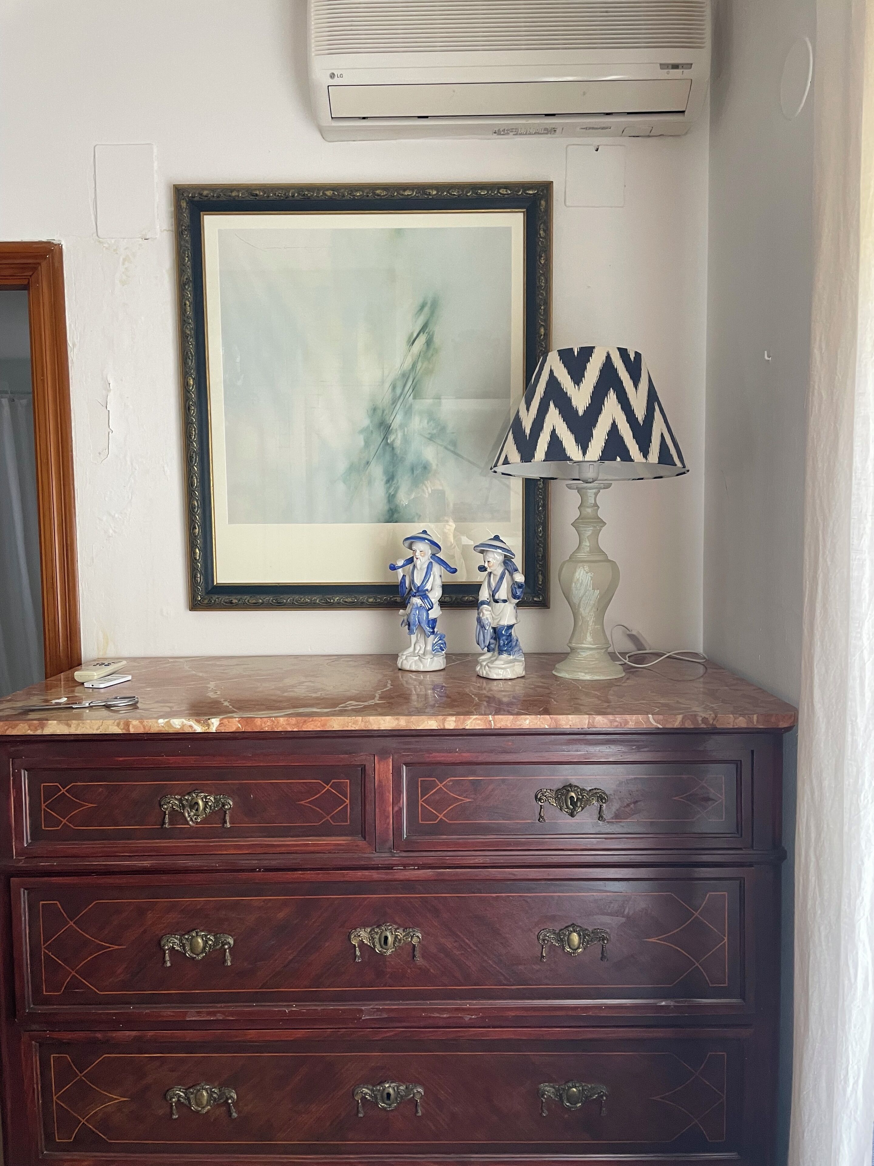 A dark wooden dresser with ornate handles, a framed abstract painting, and two ceramic figurines on top.