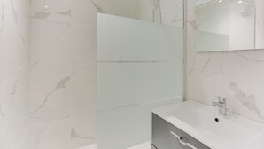 Ein modernes Badezimmer mit einer Duschkabine mit Marmorplatten, einem großen Spiegel und einem weißen Waschbecken mit einem einzelnen Wasserhahn.