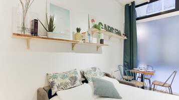 A bedroom with a bed, a small table with chairs, a shelf with decorative items, and a window with curtains.