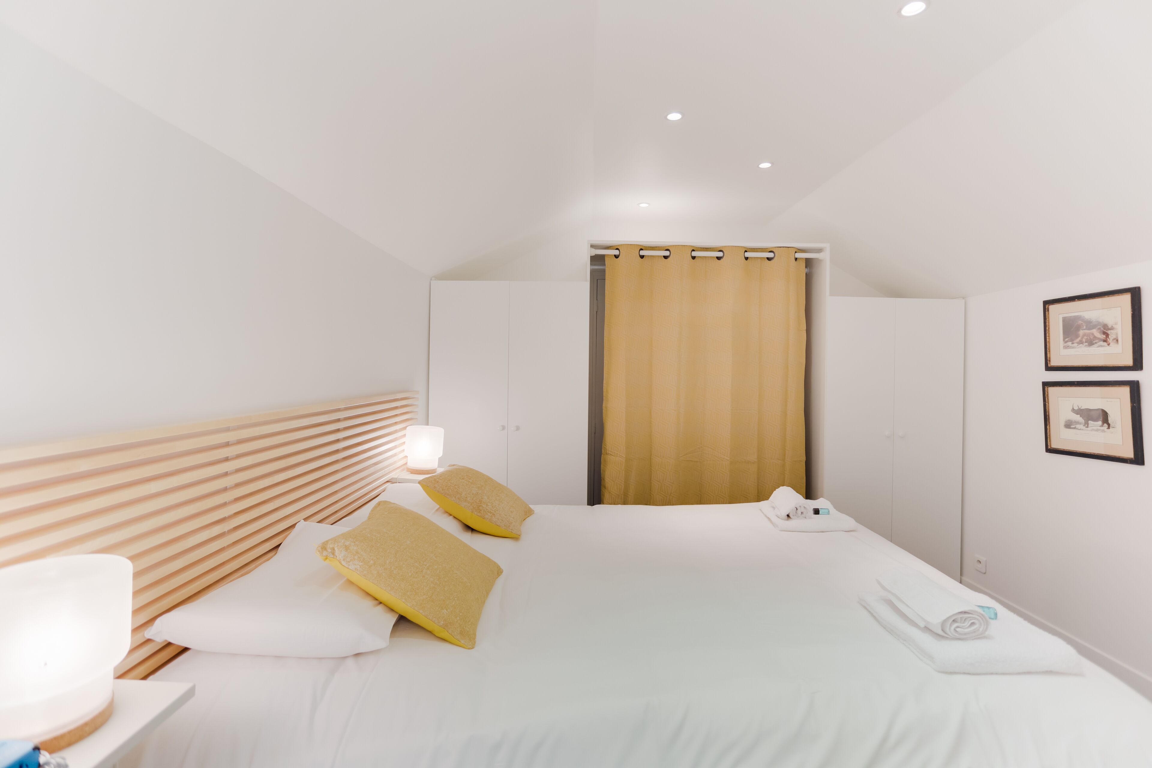 Una habitación de hotel moderna con cabecera de madera, una cama grande con ropa blanca, una mesita de noche con lámpara y una cortina.