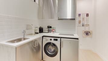 Une cuisine compacte équipée d’un lave-linge, de meubles en acier inoxydable et d’un plan de travail blanc.