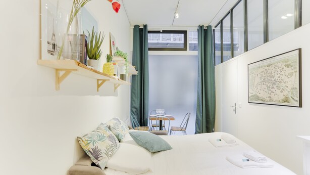 Ein modernes Hotelzimmer mit einem Bett, einem kleinen Tisch mit Stühlen, einer Ablagefläche mit Pflanzen und Büchern sowie einem Fenster mit Vorhängen.