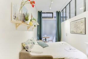 A modern hotel room with a bed, a small table with chairs, a shelf with plants and books, and a window with curtains.