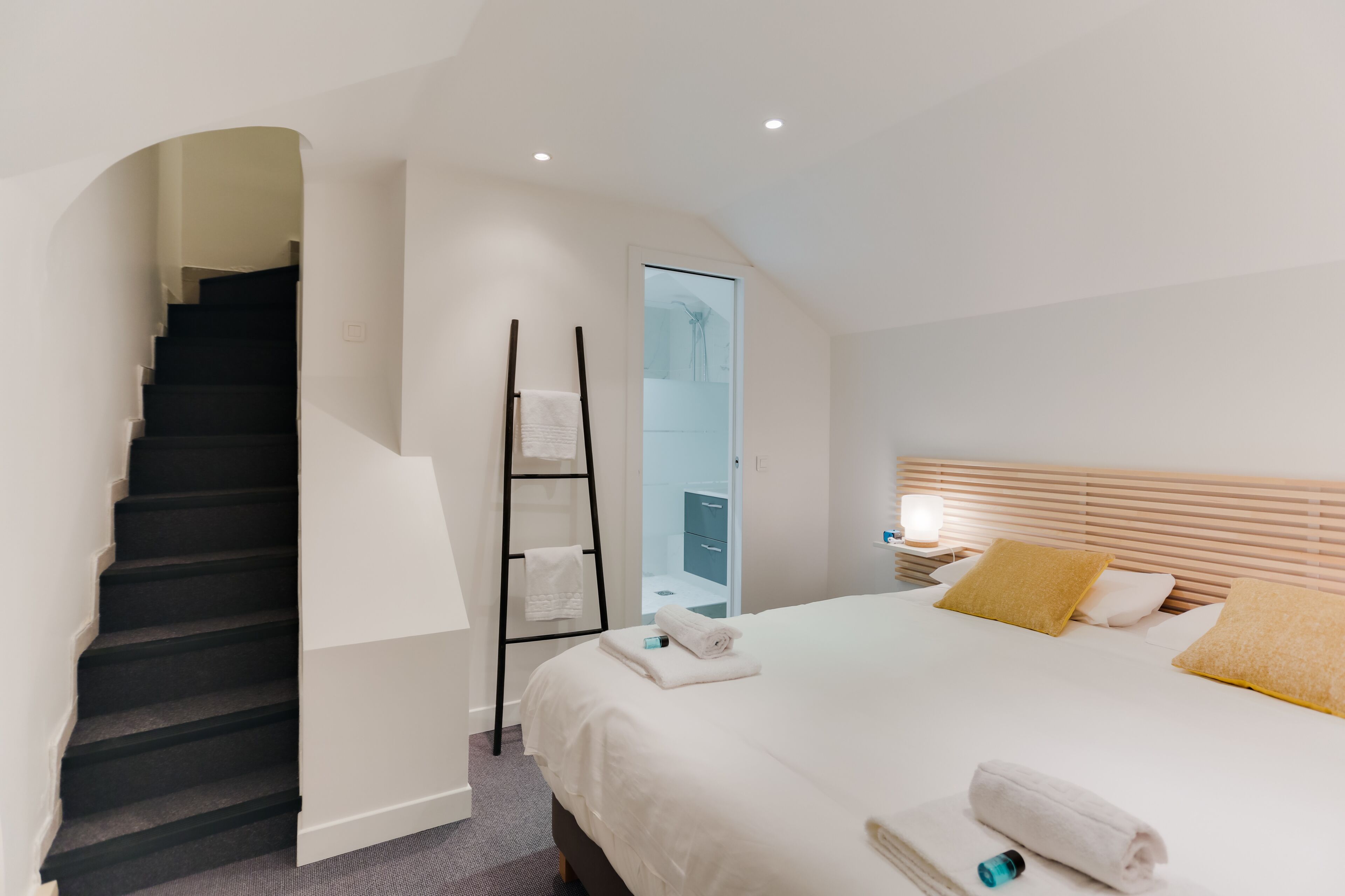 Camera d'albergo moderna con una scala, un letto con lenzuola bianche, una testiera in legno e un comodino con una lampada.