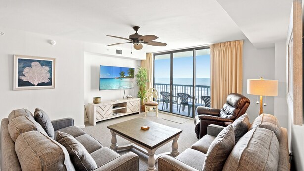 A spacious living room with a large flat-screen TV, a ceiling fan, and a view of the ocean through a sliding glass door.