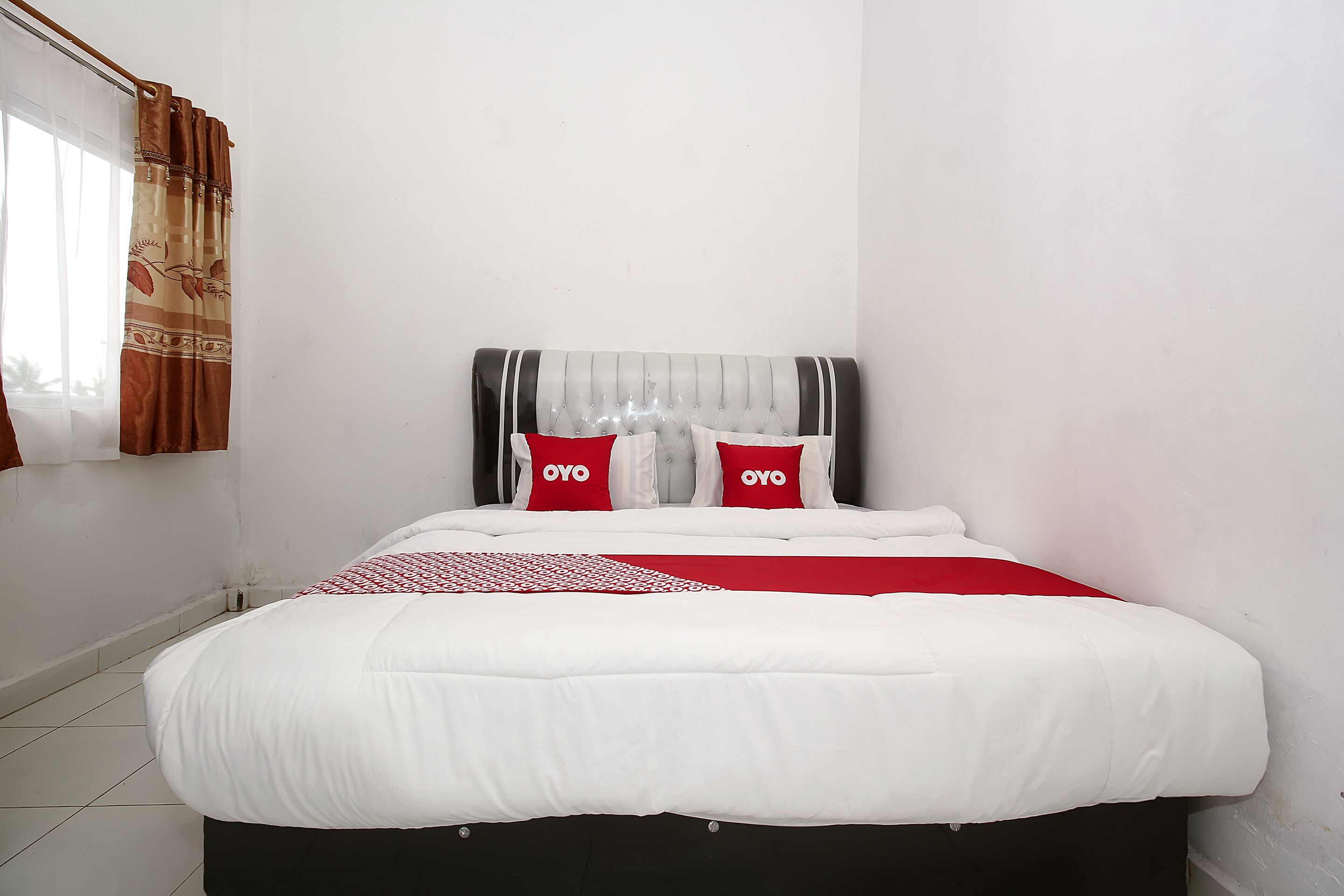 A neatly made bed with white bedding and red pillows, a grey upholstered headboard, and a red and white patterned throw.