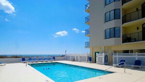 Una piscina con sillones de descanso, vista al océano y un edificio de varios pisos con balcones.