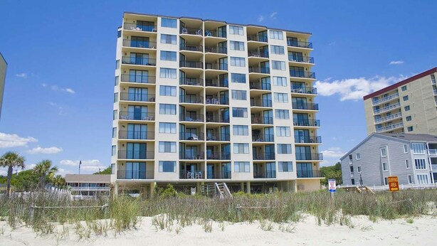 Un hotel de varios pisos con balcones, situado cerca de una playa con un cartel que indica zona restringida.