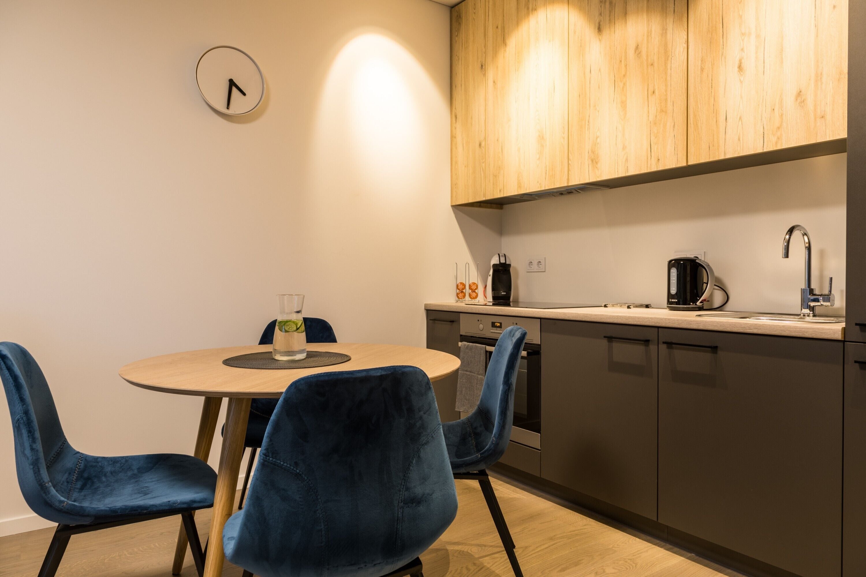 A modern kitchen with a round table, two blue chairs, wooden cabinets, and a clock on the wall.