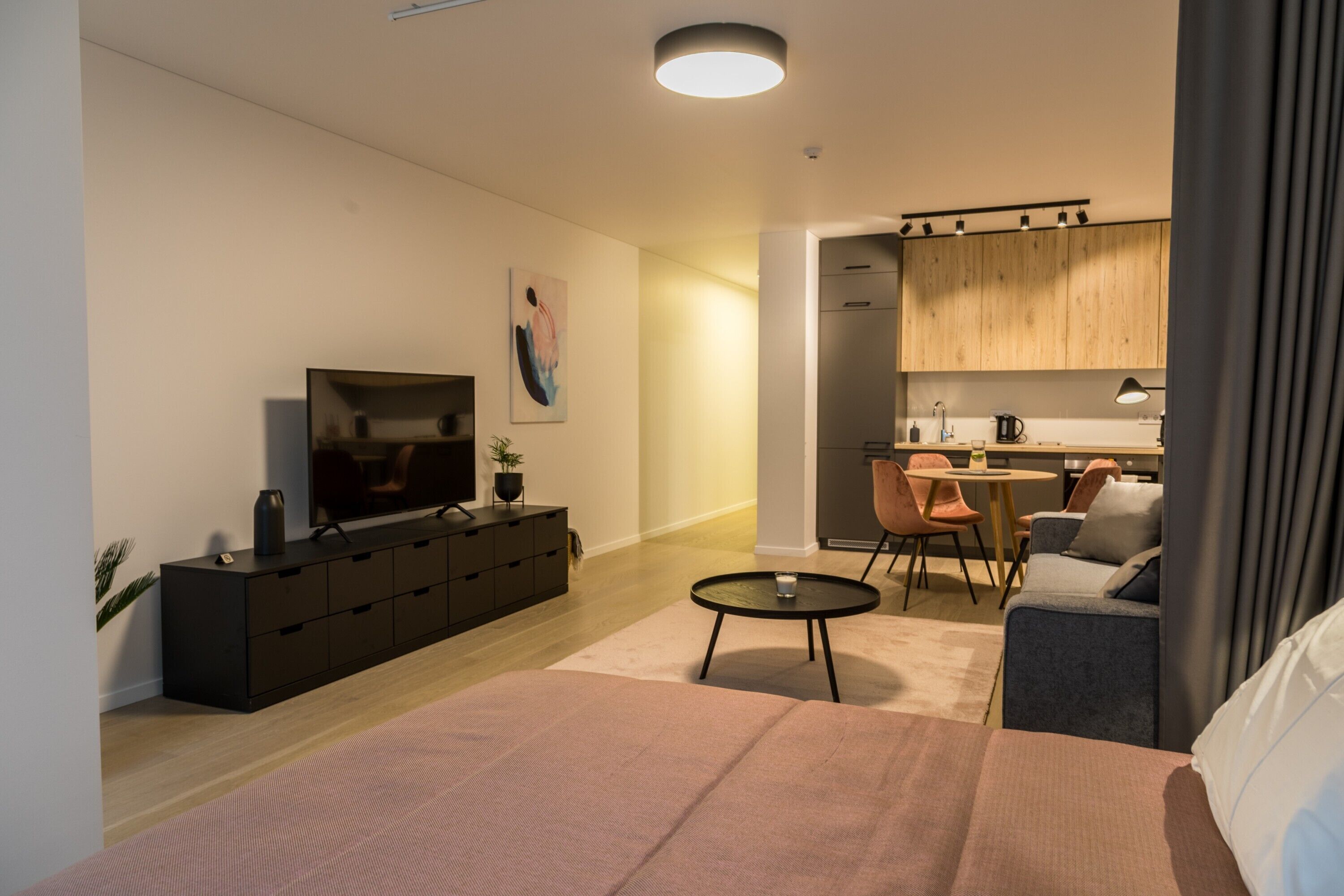 Een moderne woonkamer met een groot bed, een televisie op een zwarte kast, een klein rond tafeltje en een keuken met houten kasten en een eettafel.