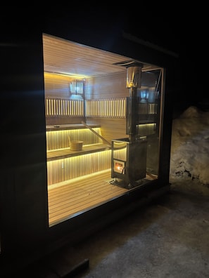 Eine Sauna mit Holzschlitzen und einer Glaseinfassung, einer Sitzbank sowie einem durch das Glas sichtbaren Heizgerät.