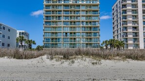 A multi-story hotel with balconies, located near a sandy beach with sparse vegetation. - Tropical Oceanfront Spacious Condo, Outdoor Hot Tub | Emerald Cove - 7C (North Myrtle Beach)