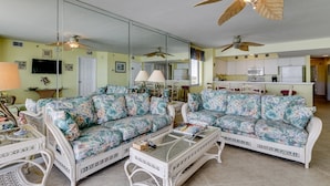 A living room with a mirrored wall, two floral sofas, a glass coffee table, and ceiling fans. - Tropical Oceanfront Spacious Condo, Outdoor Hot Tub | Emerald Cove - 7C (North Myrtle Beach)