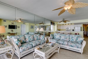 A living room with a mirrored wall, two floral sofas, a glass coffee table, and ceiling fans.