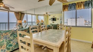 A dining area with a wooden table and chairs, a ceiling fan, and a view of the ocean through large windows. - Tropical Oceanfront Spacious Condo, Outdoor Hot Tub | Emerald Cove - 7C (North Myrtle Beach)