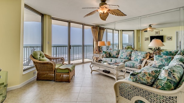 A spacious living room with a large window overlooking the ocean, a ceiling fan, and a tropical-themed sofa. - Tropical Oceanfront Spacious Condo, Outdoor Hot Tub | Emerald Cove - 7C (North Myrtle Beach)