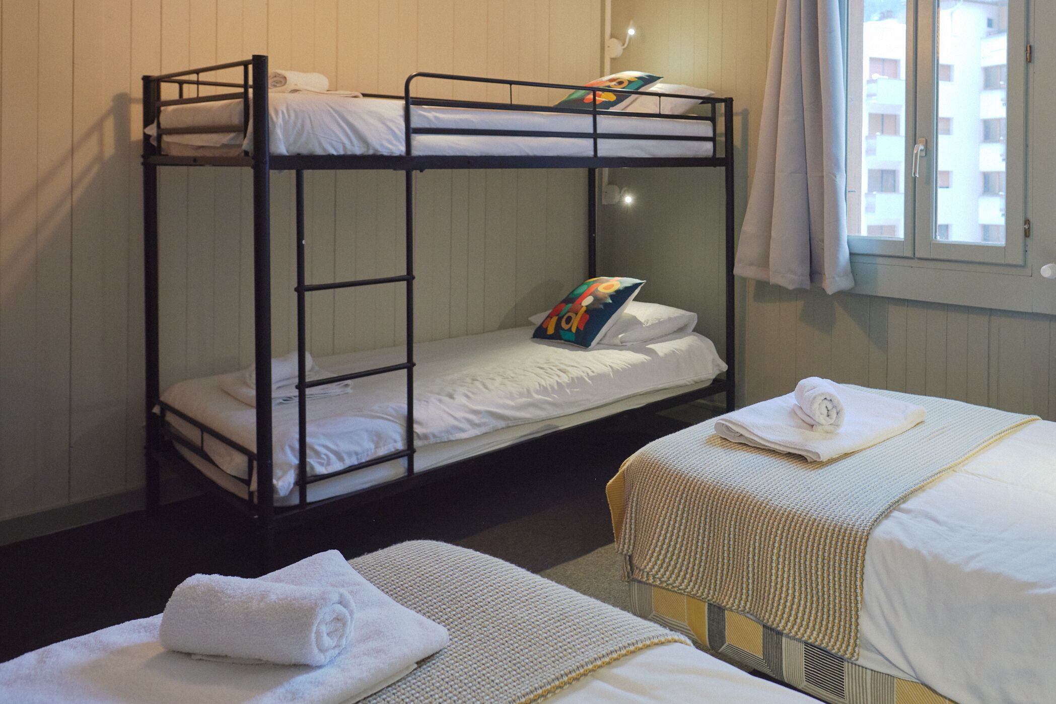 A bunk bed room with a window, a bedside table, and a small chair.