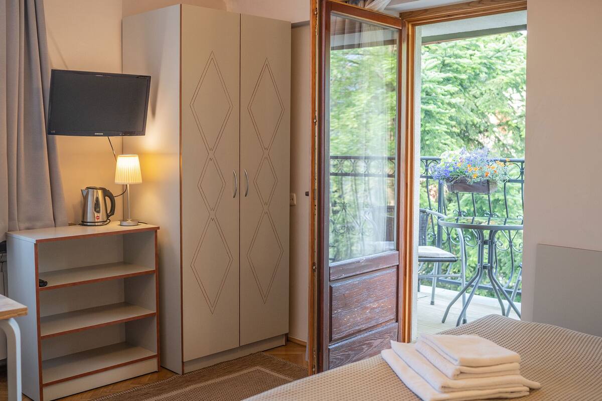A hotel room with a bed, a wardrobe, a desk with a lamp, a television, and a balcony with a chair and flowers.