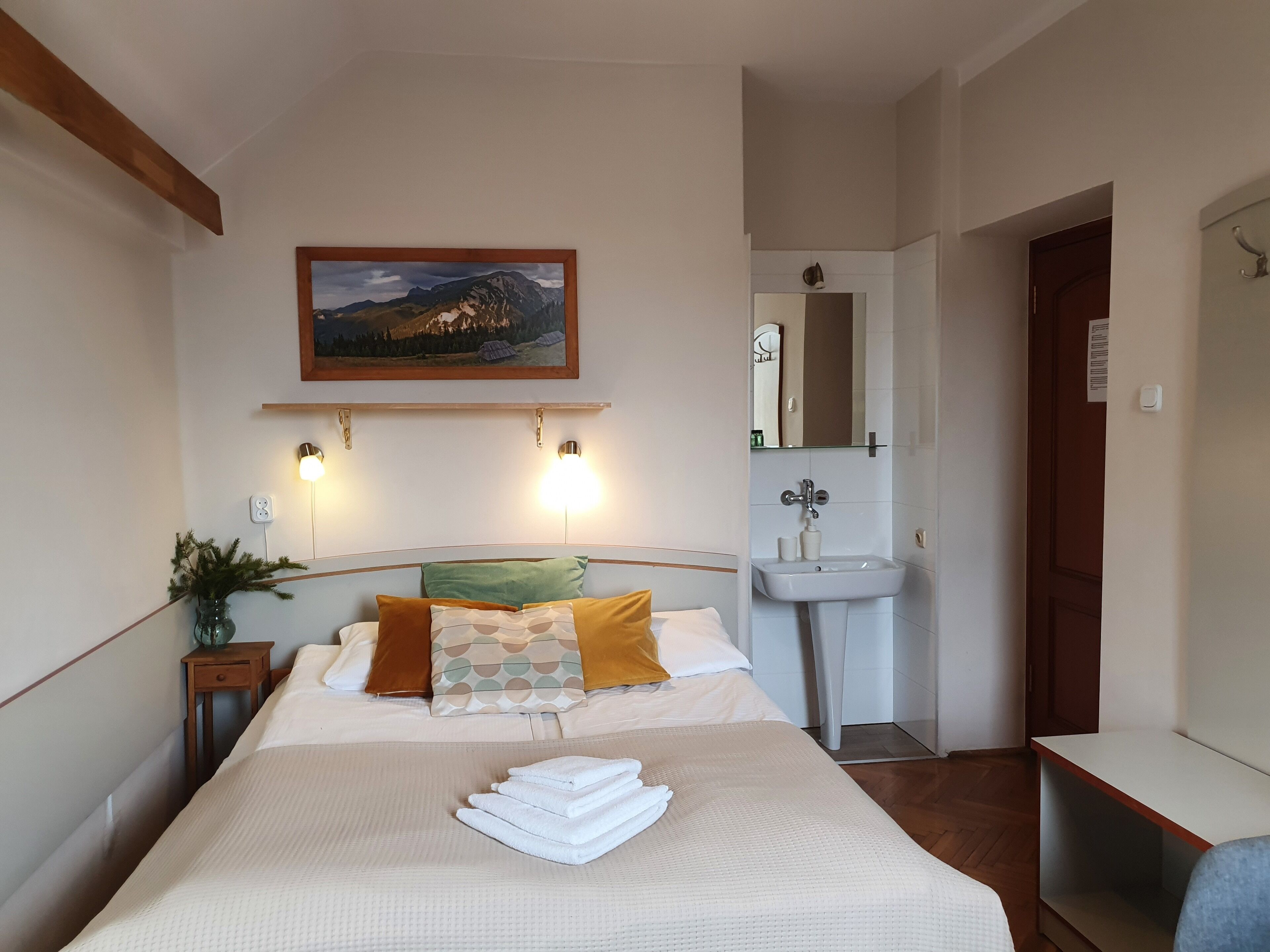 A bedroom with a bed, pillows, a bedside table with a plant, a painting on the wall, a bathroom with a sink and mirror, and a door.