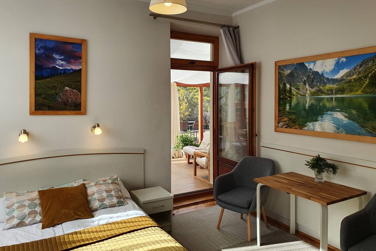A hotel room with a bed, a desk, a chair, and a view of a mountain lake through a glass door.