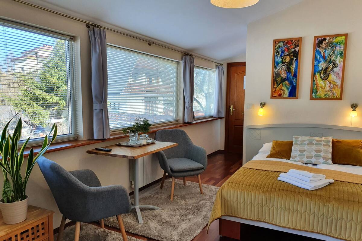 A bedroom with a bed, two chairs, a small table, a potted plant, and artwork on the wall.