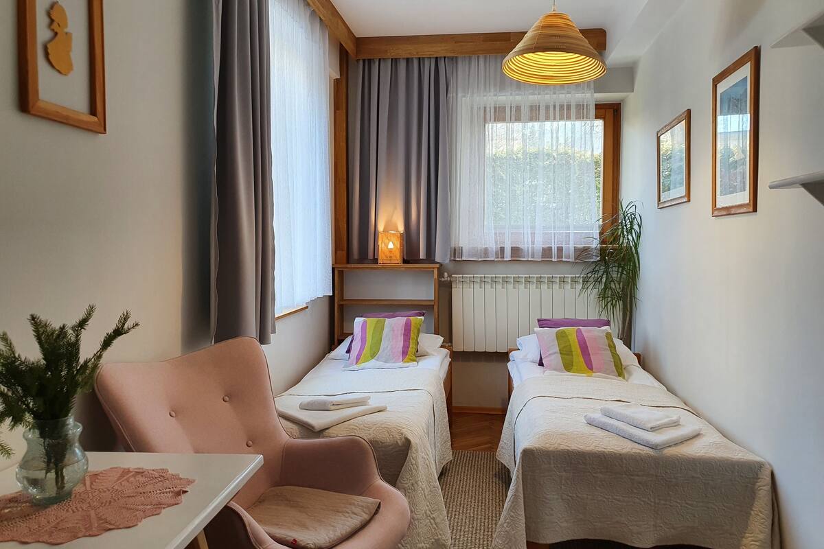 A hotel room with two single beds, a wooden headboard, a small table with a vase, a chair, and a window with curtains.
