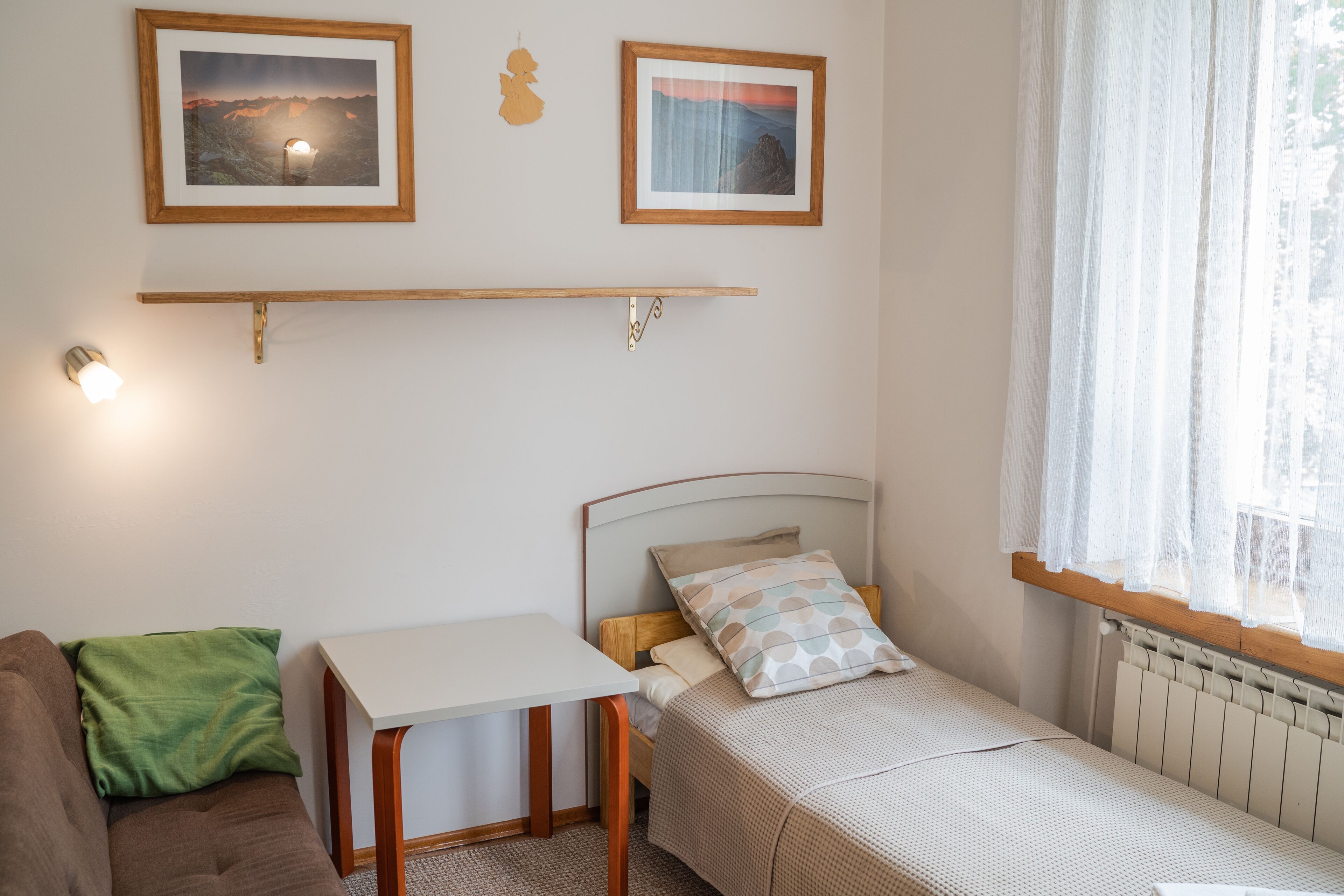 Habitació petita i acollidora amb un llit, un escriptori i un sofà. Hi ha dos quadres al mur, una finestra amb cortines primes i un radiador sota la finestra.