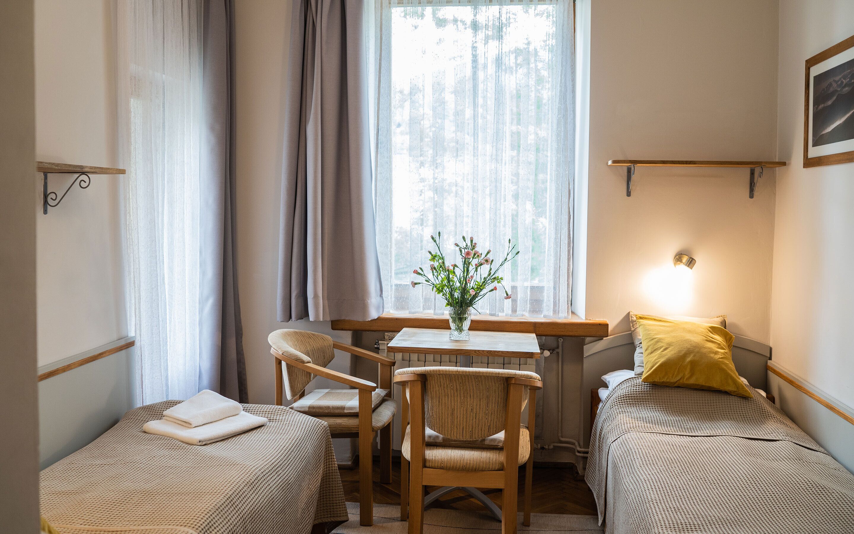 Une chambre avec deux lits simples, une chaise en bois, une petite table sur laquelle est posé un vase de fleurs, et une fenêtre avec des rideaux.