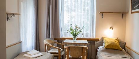 Une chambre avec deux lits simples, une chaise en bois, une petite table sur laquelle est posé un vase de fleurs, et une fenêtre avec des rideaux.