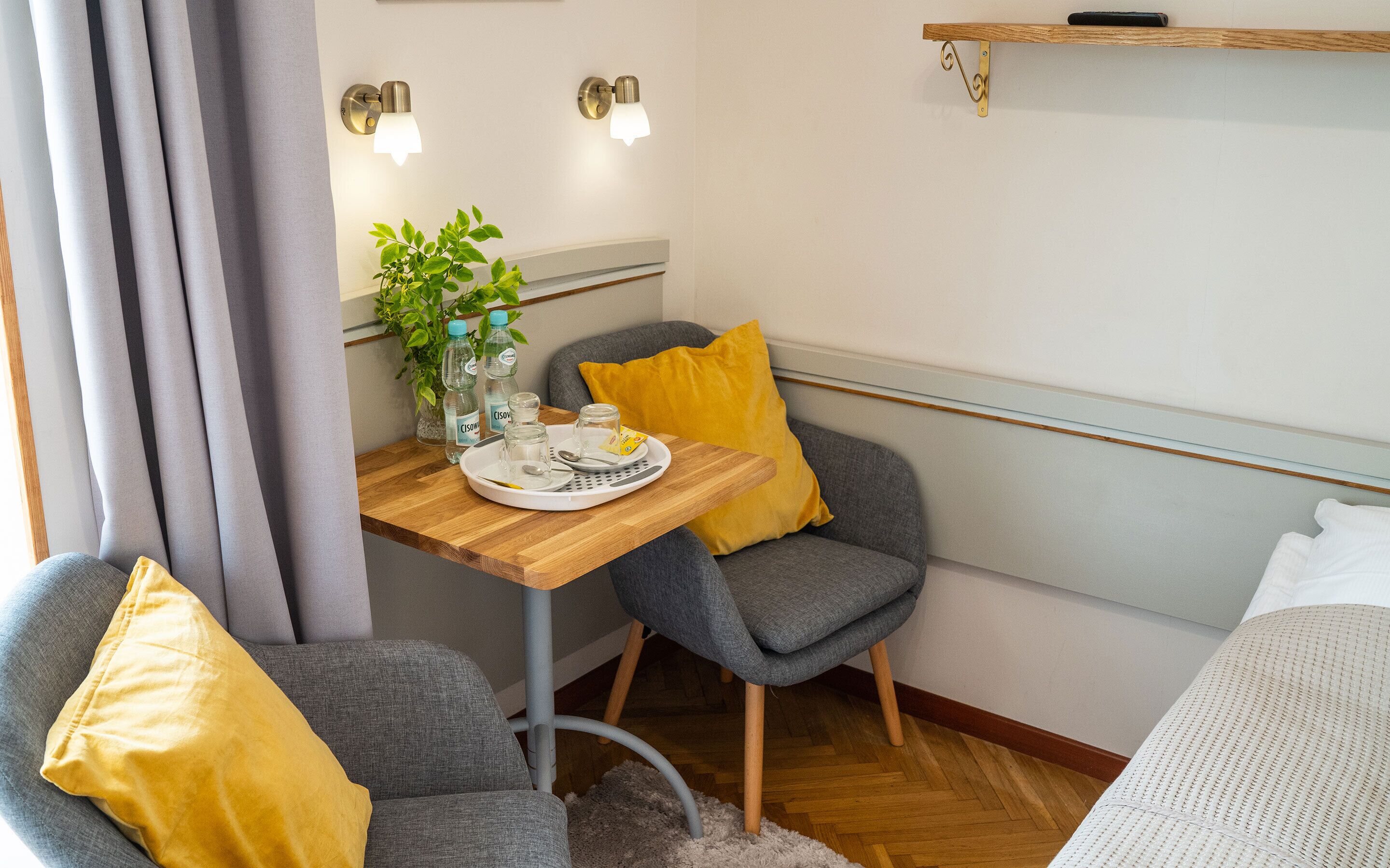 Una habitación pequeña y acogedora con una mesa de madera, dos sillas grises, un cojín amarillo y una cama blanca.