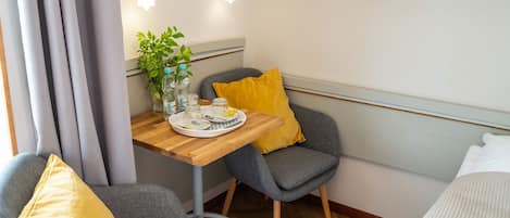 Una habitación pequeña y acogedora con una mesa de madera, dos sillas grises, un cojín amarillo y una cama blanca.