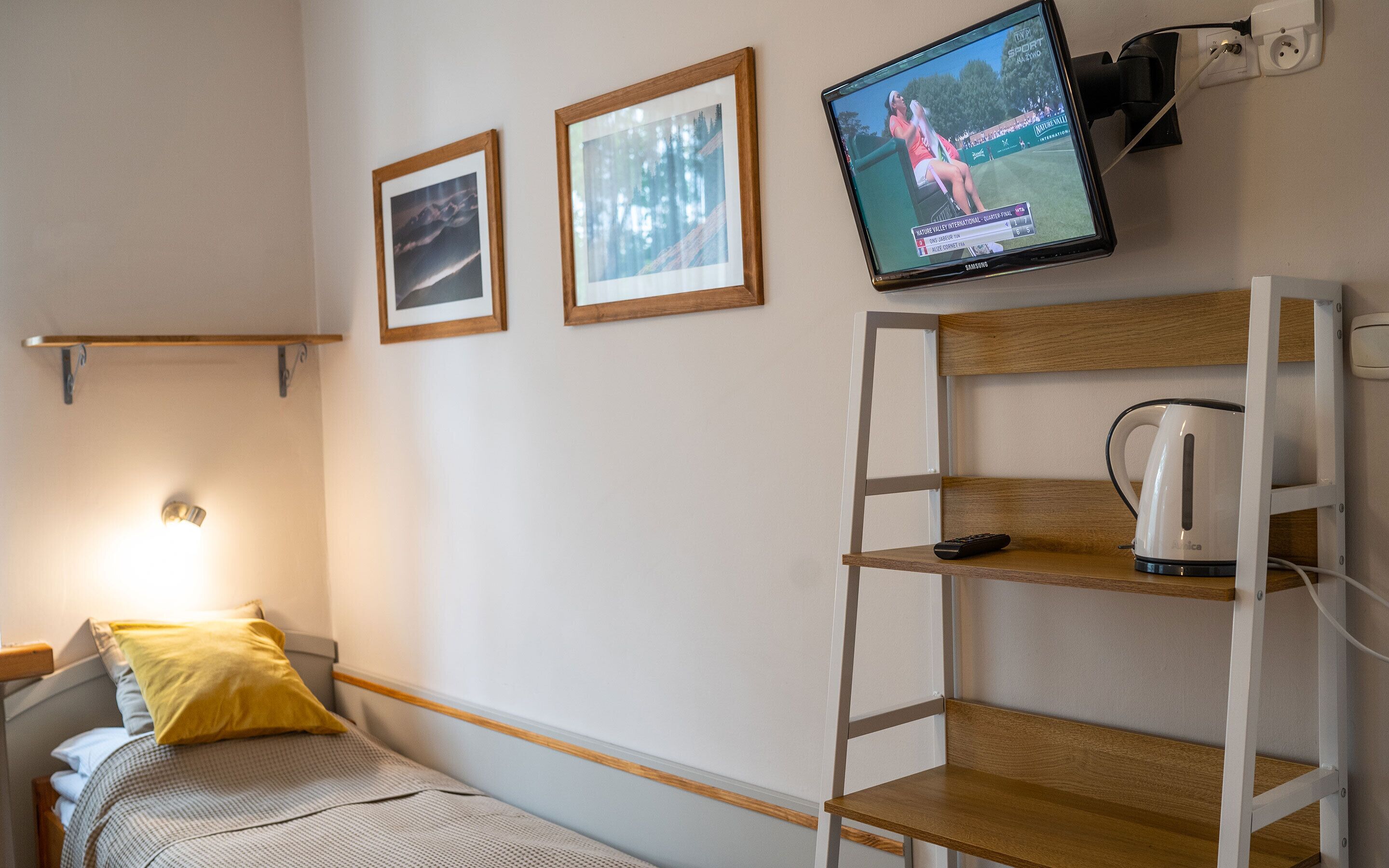 A single bed with a headboard, a bedside table with a lamp, a wall-mounted TV, and a shelf with a kettle.