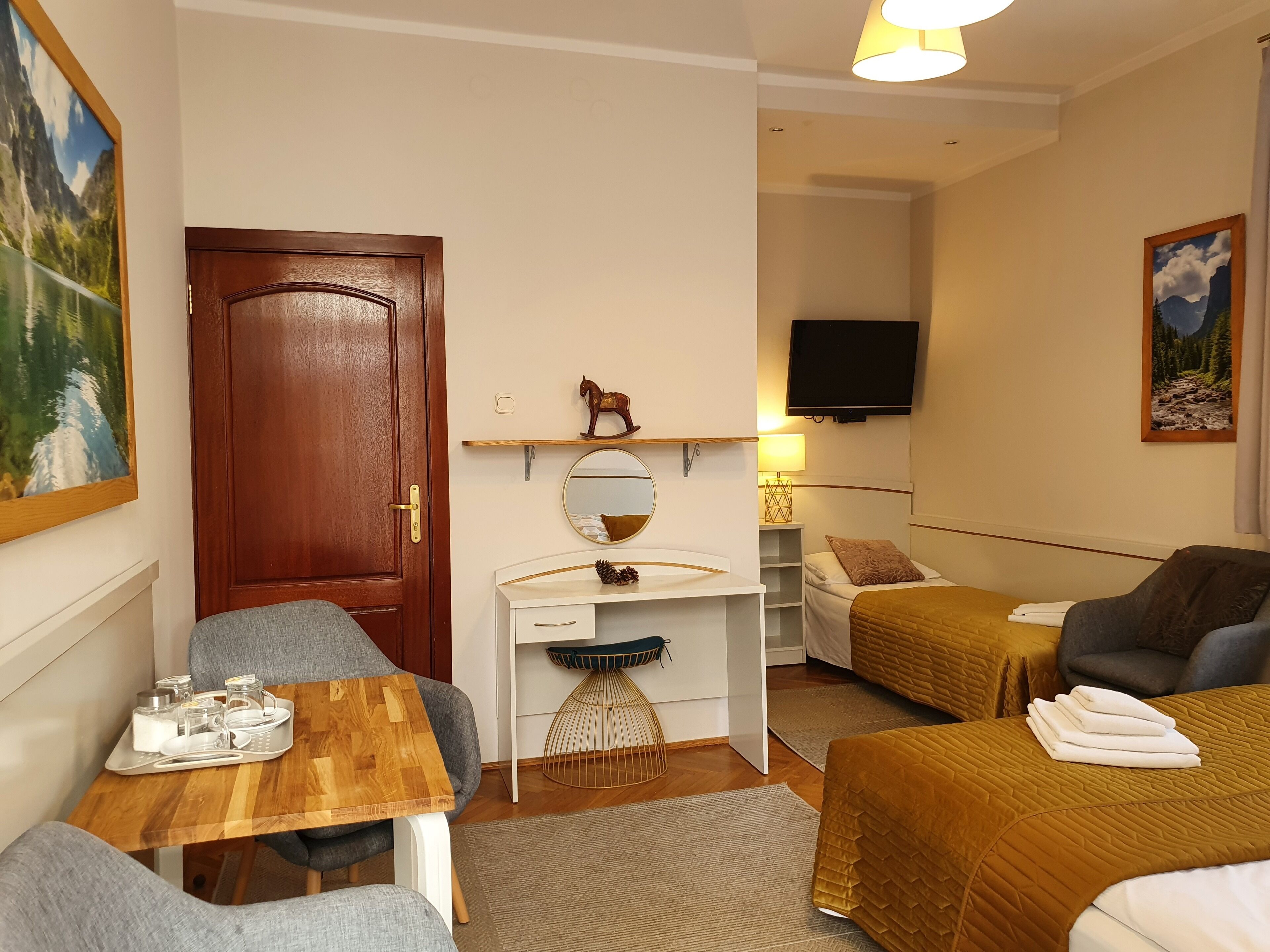 A cozy room with a wooden door, a small white desk with a mirror, a bed with a brown quilt, and a TV mounted on the wall.