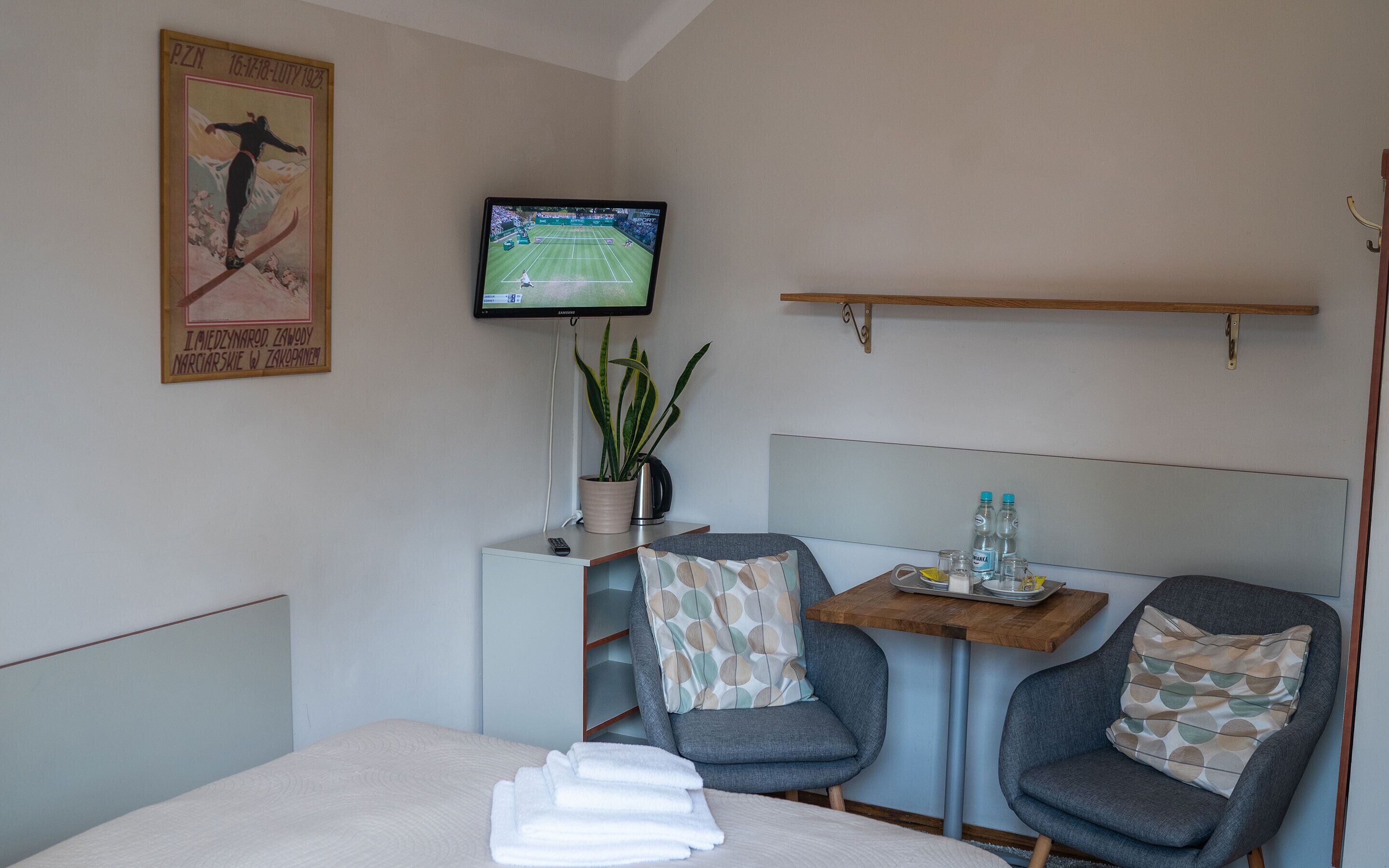 Habitación de hotel con cama, dos sillones, una mesita con una planta y botellas, un televisor en la pared y un cartel en la pared.