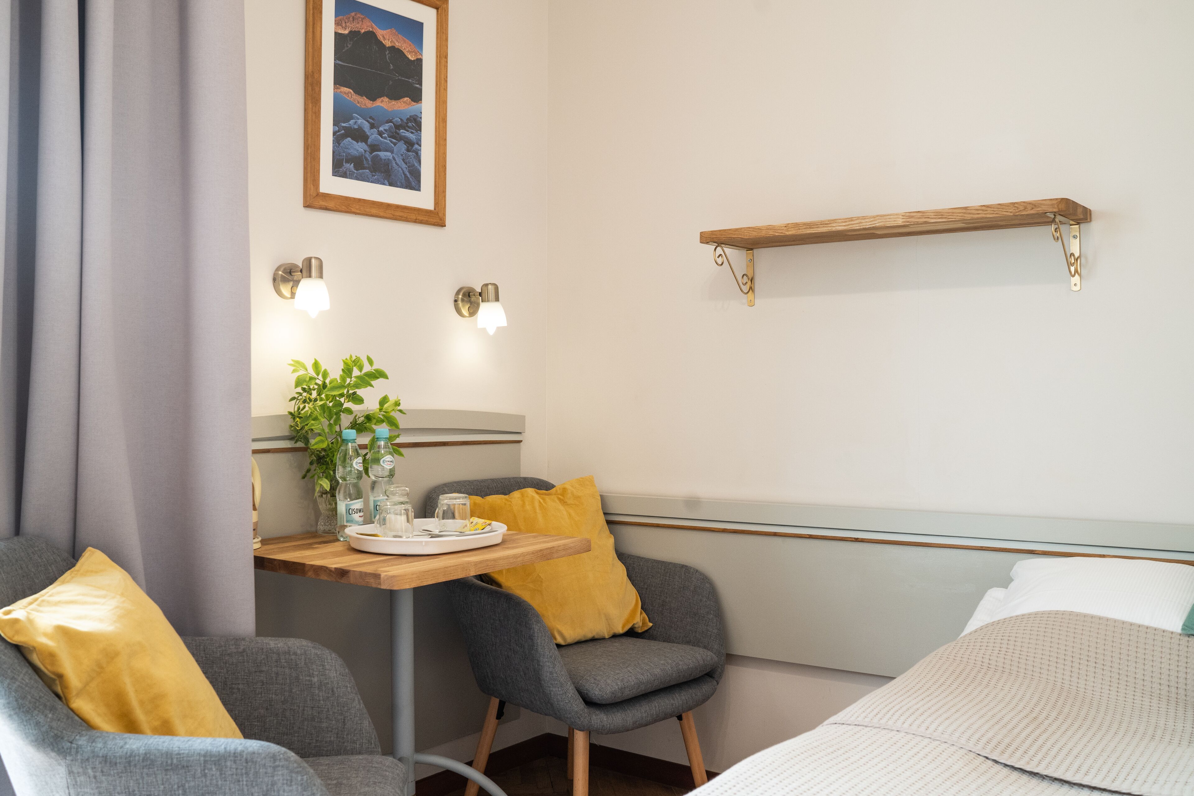 Une chambre d’hôtel comprenant un lit, deux fauteuils, une petite table avec une plante et un plateau, une étagère fixée au mur et un tableau accroché au mur.
