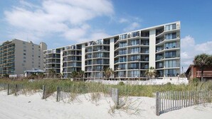 A modern multi-story hotel with balconies, located near a sandy beach with a wooden fence. - Bright Beach Condo, Oceanfront Pool, Picnic Area | Crescent Sands WH - G1 (North Myrtle Beach)