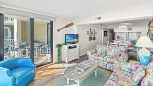 A spacious living area with a large sliding glass door leading to a balcony, a television, a patterned sofa, and a dining area with a ceiling fan. - Bright Beach Condo, Oceanfront Pool, Picnic Area | Crescent Sands WH - G1 (North Myrtle Beach)