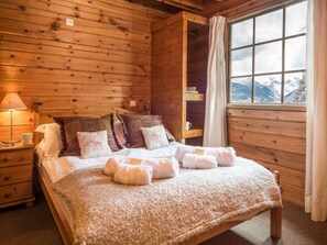 A wooden cabin bedroom with a bed, pillows, a nightstand, a window with a view of mountains, and a shelf with books.