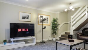 A living room with a TV displaying 'NETFLIX', a potted plant, a staircase, and a leather sofa. - Tudors eSuites Four Bedroom House (Birmingham)