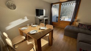 Ein Essbereich mit einem Holztisch, der für vier Personen gedeckt ist, einem Fernseher auf einem Standfuß und einem Sofa im angrenzenden Wohnbereich.