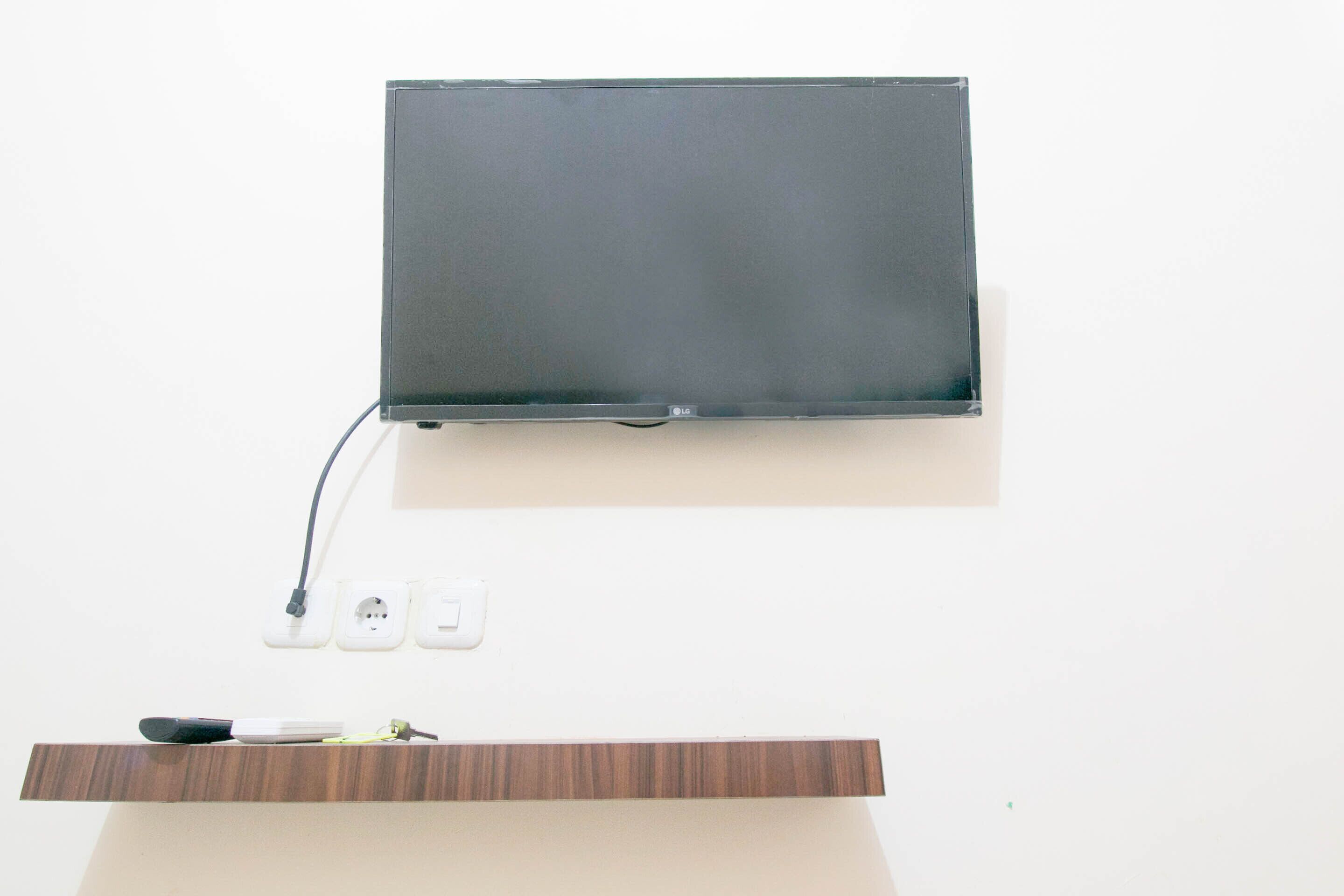 Un televisor de pantalla plana montado en una pared blanca, con un enchufe y un estante de madera debajo.