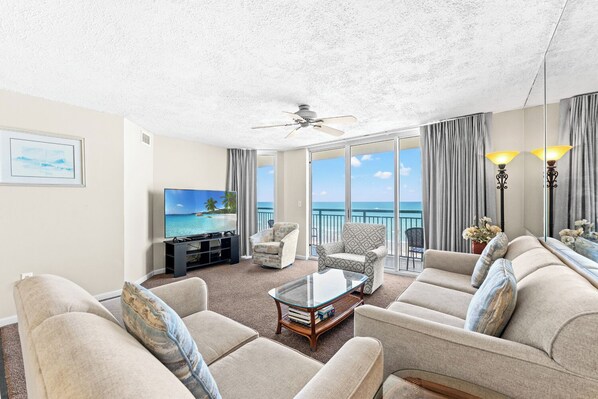 A living room with a television, seating, a glass coffee table, and a view of the ocean.