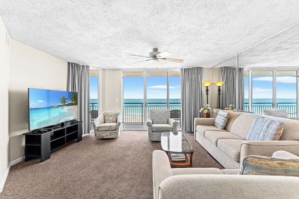 A living room with a television, two armchairs, a sofa, a glass coffee table, and a ceiling fan.