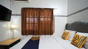A hotel room with a bed, a TV mounted on the wall, a bedside table with a lamp, and a window with curtains. - SPOT ON 1854 Sepinggan Asri Syariah Guesthouse (Balikpapan)