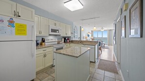 A modern kitchen with white cabinets, a granite countertop, and a central island. There is a microwave above the stove, and a refrigerator on the left. The kitchen has a large window providing natural light. - Nautical Oceanfront Condo, Outdoor Covered Heated Pool | Pinnacle - 802 (North Myrtle Beach)