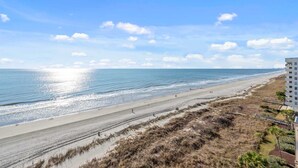 A beach with a sandy shore, a clear sky, and a building on the right. - Nautical Oceanfront Condo, Outdoor Covered Heated Pool | Pinnacle - 802 (North Myrtle Beach)