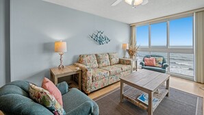 A living room with a sofa, two armchairs, a coffee table, and a side table with lamps. There is a large window with a view of the ocean. - Nautical Oceanfront Condo, Outdoor Covered Heated Pool | Pinnacle - 802 (North Myrtle Beach)
