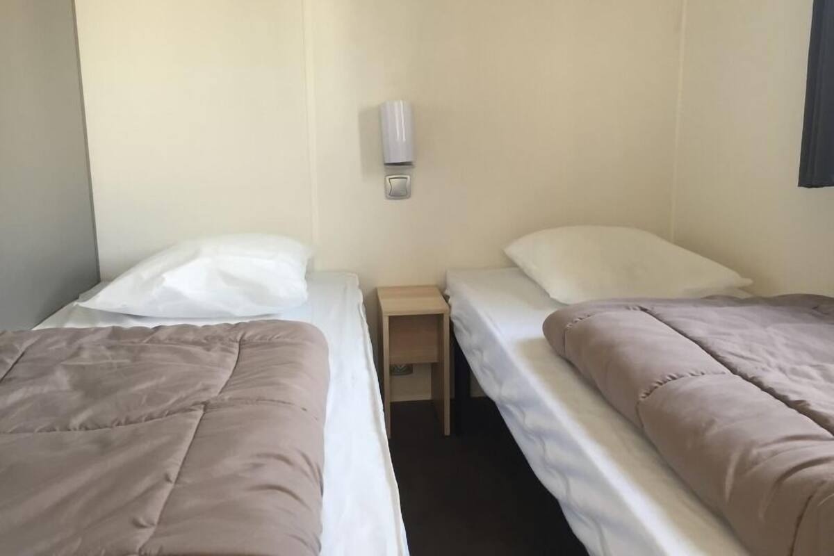 A small, simple room with two beds, each with a pillow and a blanket. There is a small wooden nightstand between the beds.