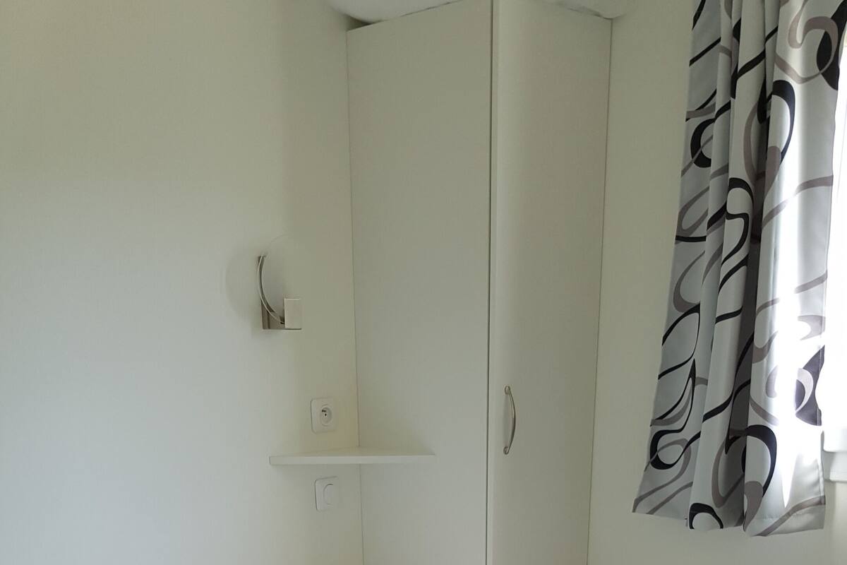 A single bed with a white sheet and a brown blanket, a white wardrobe, a small shelf, and a window with patterned curtains.